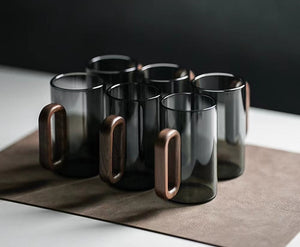 Two Fiorella Smoky Fashioned Drinking Glasses by Macchiaco, each 11oz with sleek wooden handles, are displayed on a brown mat atop a white surface against a dark background.
