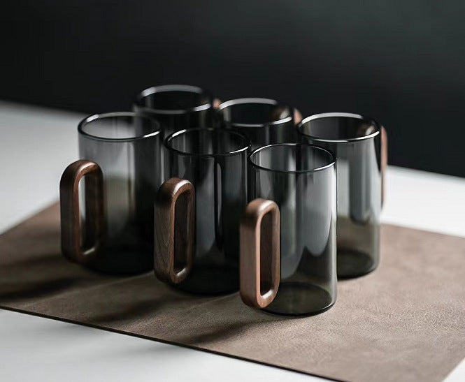 Two Fiorella Smoky Fashioned Drinking Glasses by Macchiaco, each 11oz with sleek wooden handles, are displayed on a brown mat atop a white surface against a dark background.