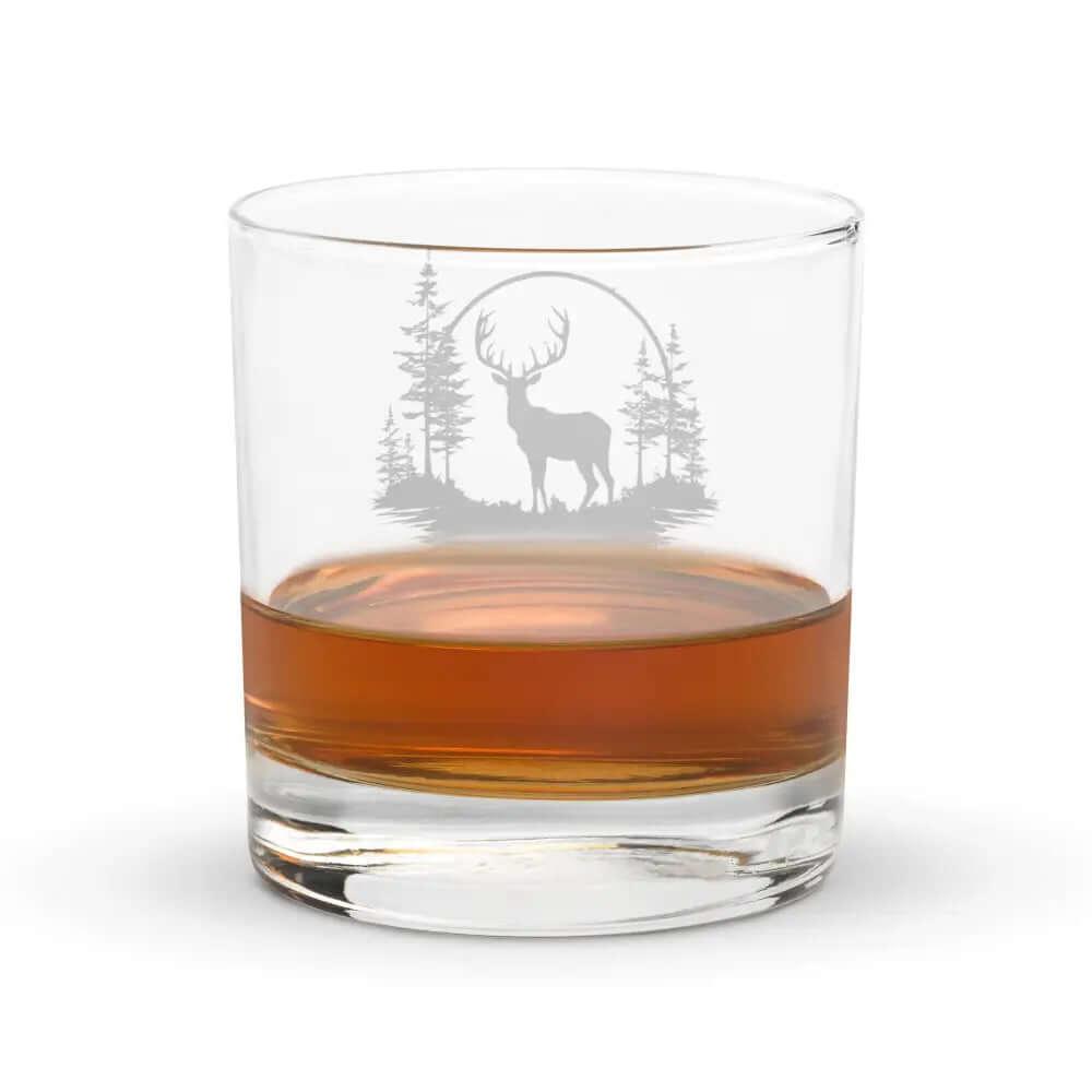 The 10.5 oz Deer Treeview Rocks Glass is a whiskey or bourbon glass featuring an etched design of a deer standing among trees within a circular frame, shown holding a brown liquid.