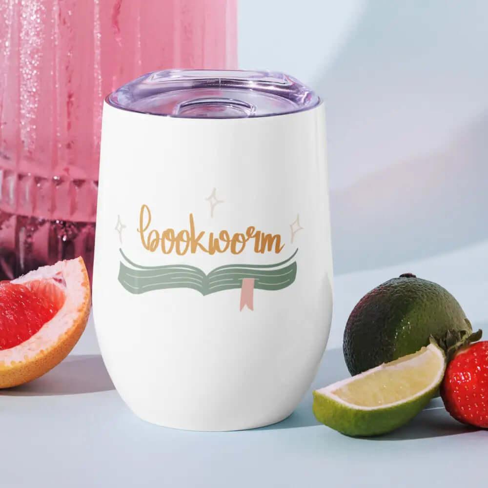 The 12 oz Bookworm Wine Tumbler, Travel Mug is a white stainless steel cup with bookworm above a book graphic and clear lid—ideal outdoor party drinkware for literary fans, styled beside sliced grapefruit, lime, and a strawberry.