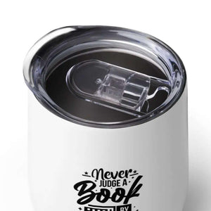 A 12 oz Never Judge A Book Wine Tumbler, featuring a white double-wall vacuum seal and a clear plastic lid, is filled with a dark beverage.