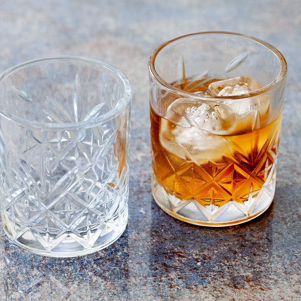 Two 7 Ounce Etched Whiskey Glasses from the set of 6 rest on a stone surface; one is empty, while the other holds an amber drink with ice cubes.