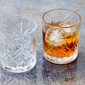 Two 7 Ounce Etched Whiskey Glasses from the set of 6 rest on a stone surface; one is empty, while the other holds an amber drink with ice cubes.
