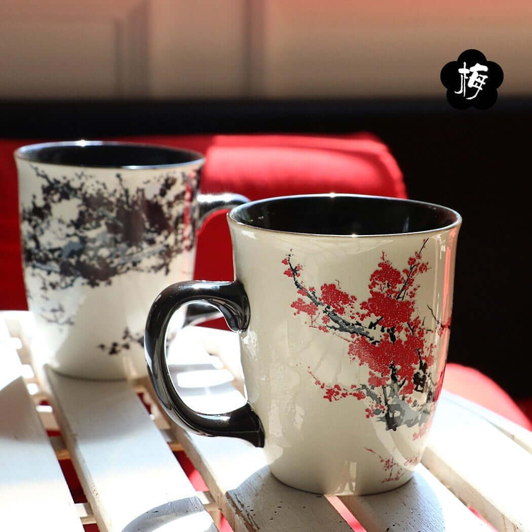 Two 16oz Color Changing Heat Sensitive Magic Porcelain Mugs, featuring black handles and floral branch designs (one with red flowers, one with black), sit on a white slatted surface with a red cushion in the background.