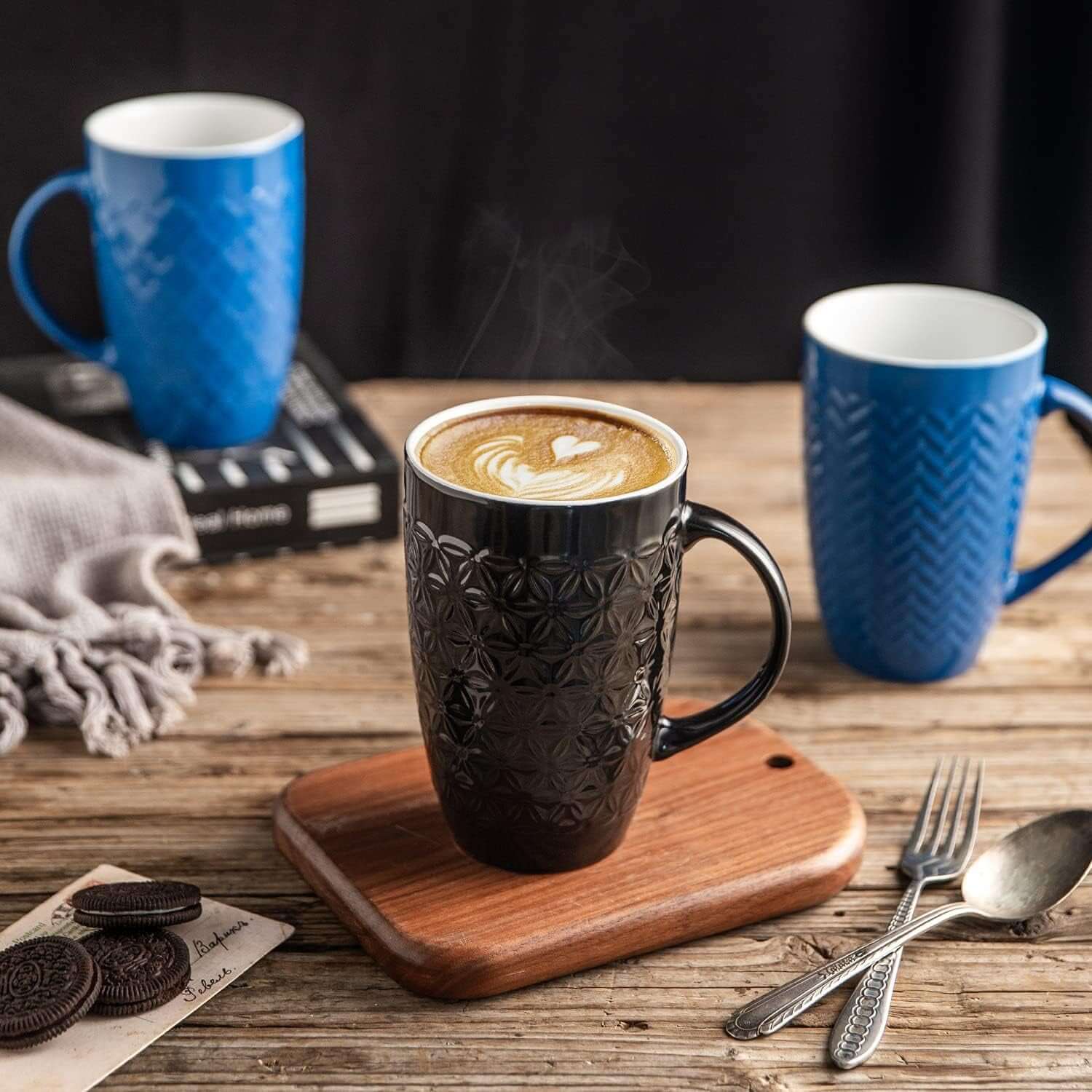 A Large Coffee Mugs Set of 6, 22oz ceramic tall coffee mugs, lead and cadmium free, is displayed on a rustic wooden table with cookies, cutlery, and a gray cloth beside them.