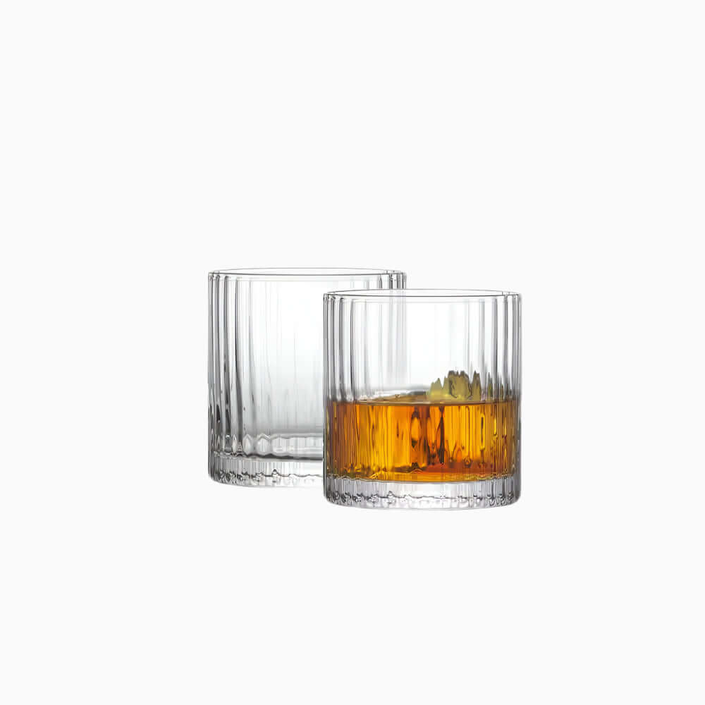 A set of two Elle Fluted Double Old Fashion Whiskey Glasses (10 oz.) sits on a white background; one glass is empty, the other holds amber whiskey with ice and a lemon twist.