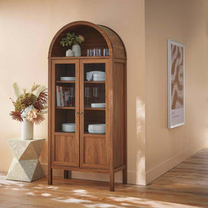 A wooden display cabinet with glass doors stands against a beige wall, holding books, dishes, and an Elle Fluted Double Old Fashion Whiskey Glass set. Nearby, a geometric side table has a vase of dried flowers, with wall art in the background.