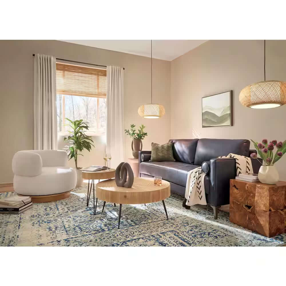 A modern living room with a black sofa, white armchair, round wooden coffee table topped with Elle Fluted Double Old Fashion Whiskey Glasses, pendant lights, decorative plants on a patterned rug, and sunlight streaming through curtained windows.