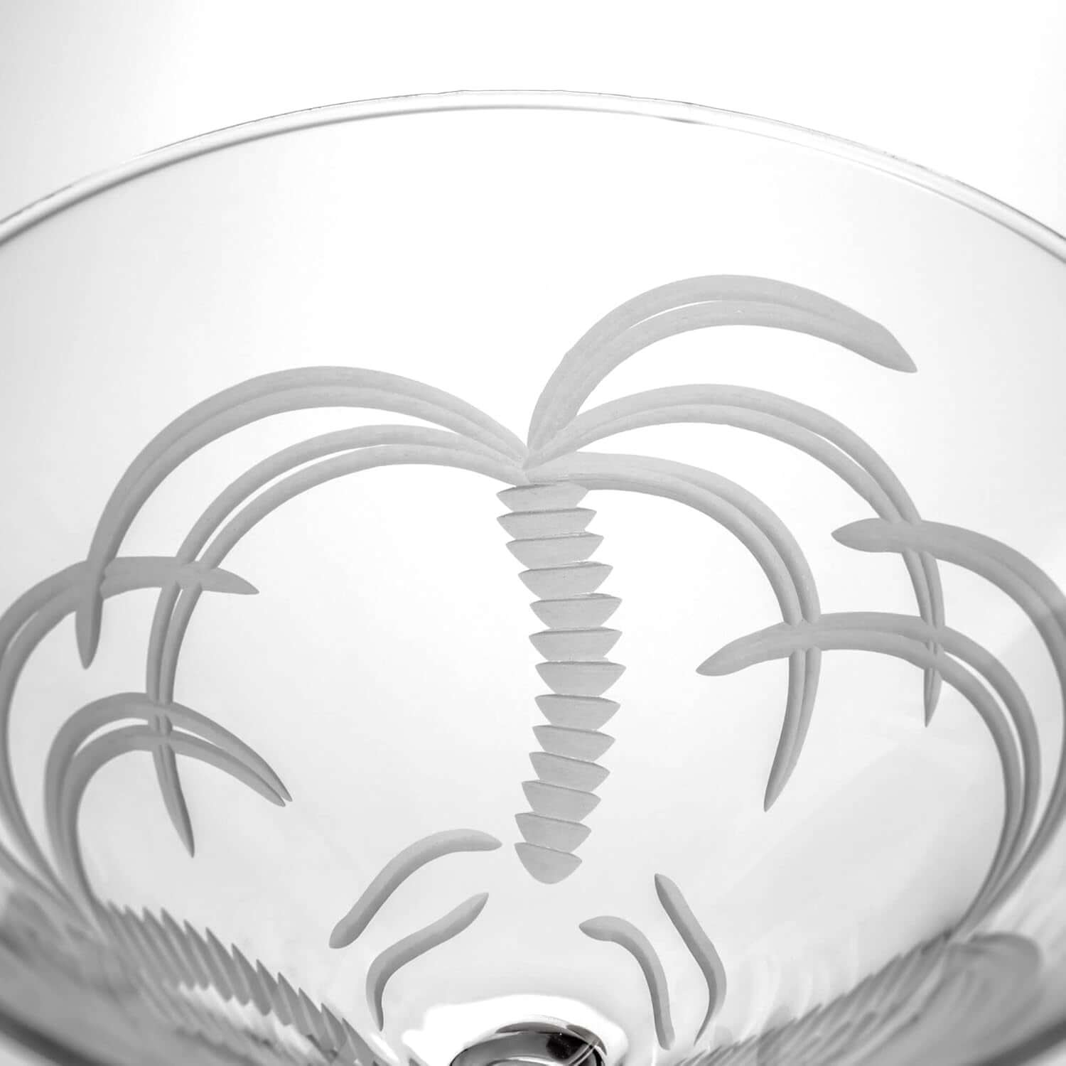 A close-up of the Palm Tree Martini Glass, a 10 oz stemmed, lead-free glass with an etched palm tree near the rim, shown against a white background. Available as a set of 4.