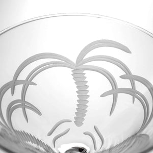 A close-up of the Palm Tree Martini Glass, a 10 oz stemmed, lead-free glass with an etched palm tree near the rim, shown against a white background. Available as a set of 4.