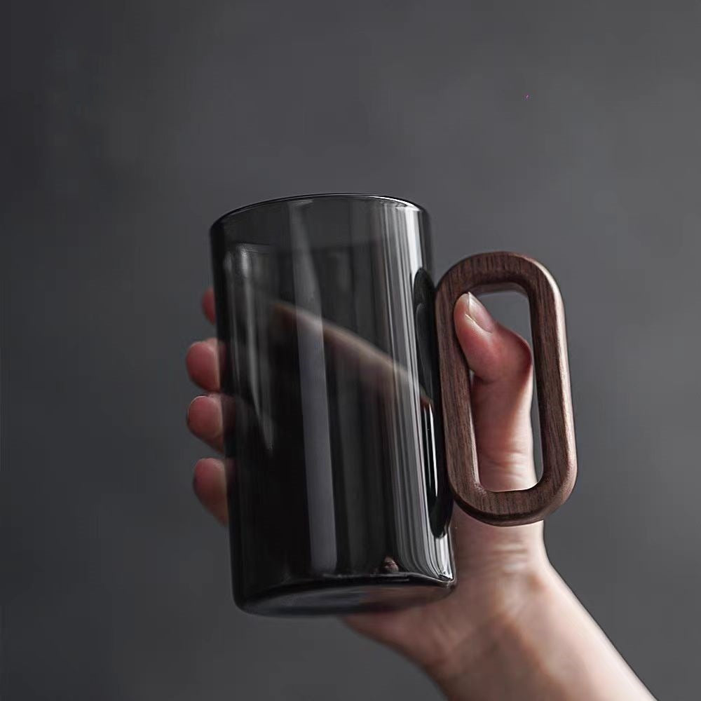 A hand holds a Macchiaco Fiorella Smoky Fashioned Drinking Glass (11oz, set of 2), featuring a large wooden handle, against a plain gray background.