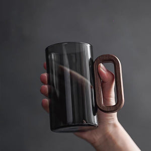 A hand holds a Macchiaco Fiorella Smoky Fashioned Drinking Glass (11oz, set of 2), featuring a large wooden handle, against a plain gray background.