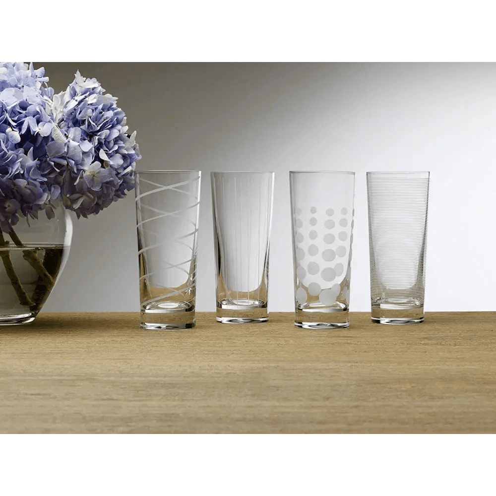 A set of four Cheers Highball Glasses, 19.75-oz each, featuring different etched patterns, is arranged in a row on a wooden surface beside a vase of purple hydrangeas.