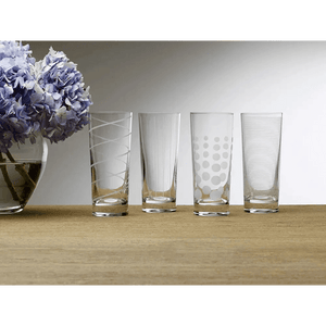 A set of four Cheers Highball Glasses, 19.75-oz each, featuring different etched patterns, is arranged in a row on a wooden surface beside a vase of purple hydrangeas.