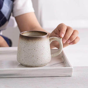 A person holds the handle of a Macchiaco 11 Oz Cocoa Coffee Mug with a speckled ceramic finish, resting it on a rectangular tray.