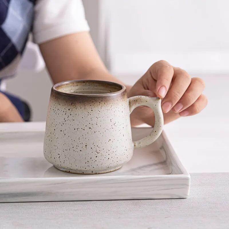 A person holds the handle of a Macchiaco 11 Oz Cocoa Coffee Mug with a speckled ceramic finish, resting it on a rectangular tray.