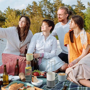 Four people enjoy a picnic outdoors, surrounded by food, drinks, and Coffee Mugs Stackable Porcelain Coffee Cups Gift Set, 15 Oz. Trees fill the background as one person takes a selfie.