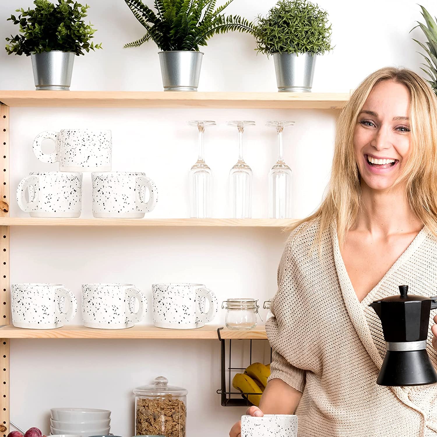 A woman stands in a kitchen holding a stovetop espresso maker and a 10 oz Ceramic Coffee Mug from the Stoneware Coffee Cups Set of 4, with shelves behind her displaying mugs, jars, plants, and fruit.