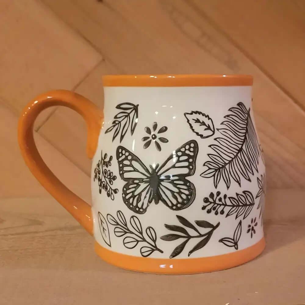 The Homey Butterfly Ceramic Coffee Mug 16 Oz features an orange handle and rim, plus black and white butterfly and leaf designs—making it the perfect autumn mug for your favorite fall coffee.