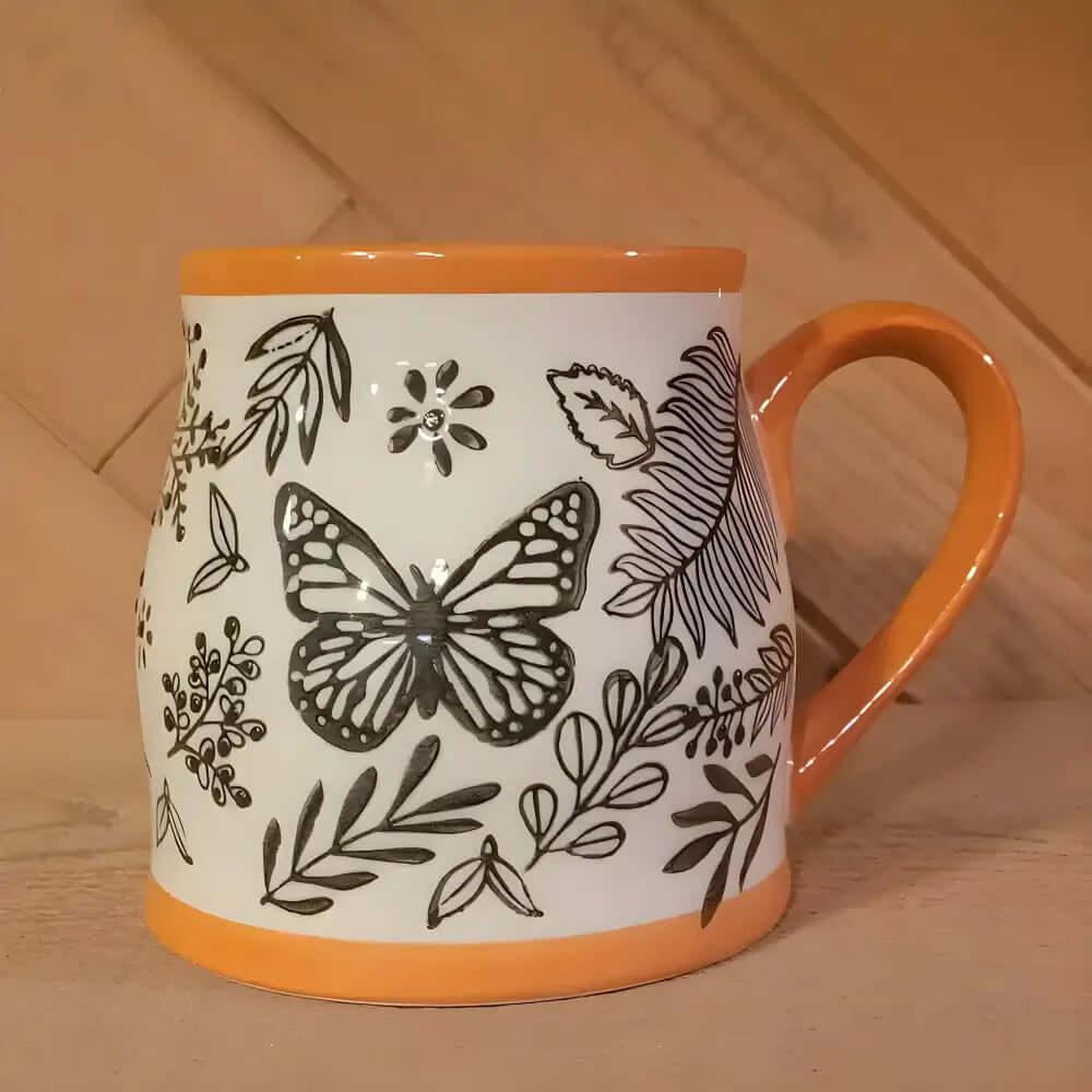 The Homey Butterfly Ceramic Coffee Mug 16 Oz has an orange rim and handle, adorned with black and white butterfly designs among leaves and flowers—an ideal autumn mug for your favorite cozy drinks.