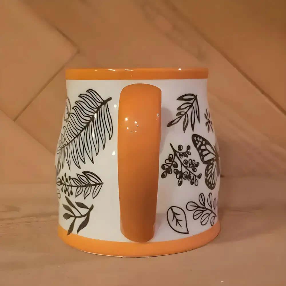 The Homey Butterfly Ceramic Coffee Mug 16 Oz features black botanical and butterfly designs with an orange handle and rim, bringing cozy autumn vibes to your coffee moments.