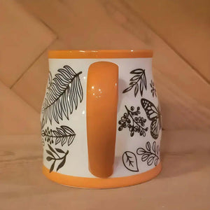 The Homey Butterfly Ceramic Coffee Mug 16 Oz features black botanical and butterfly designs with an orange handle and rim, bringing cozy autumn vibes to your coffee moments.