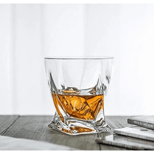 A Premium Crystal Whiskey Glasses Set of 6 Twist Large 10 oz, made from lead-free crystal, holds whiskey and ice on a wooden surface beside books or magazines.