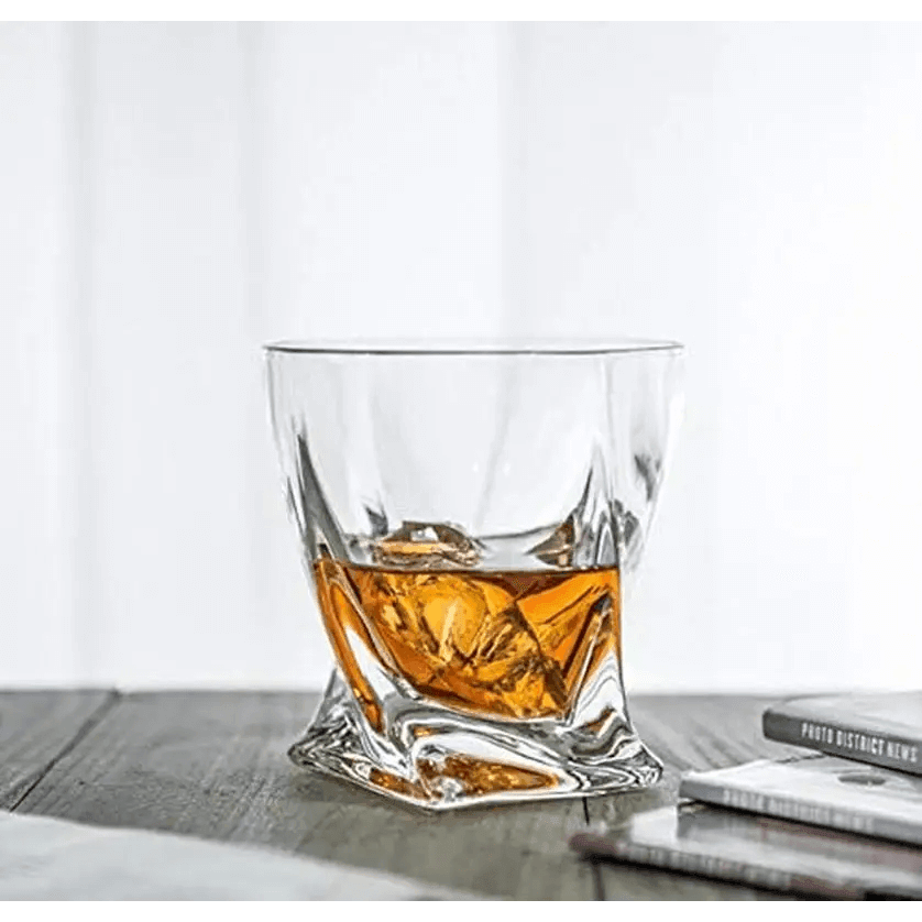 A Premium Crystal Whiskey Glasses Set of 6 Twist Large 10 oz, made from lead-free crystal, holds whiskey and ice on a wooden surface beside books or magazines.