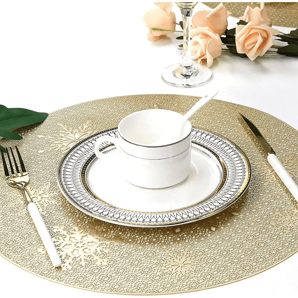 A formal table set with Round Gold Christmas placemats (Set of 4), white and silver plates, a cup and spoon, white-handled cutlery, a wine glass, and peach roses in the background.