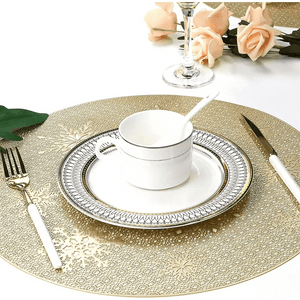 A formal table set with Round Gold Christmas placemats (Set of 4), white and silver plates, a cup and spoon, white-handled cutlery, a wine glass, and peach roses in the background.