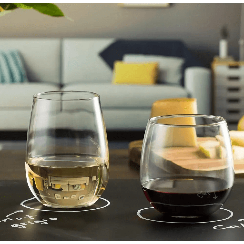 Two glasses from the Stemless 12-Piece Wine Glass Party Set (16.75 oz, 17 oz), one with white and one with red wine, rest on a black tabletop in a modern living room featuring a sofa and colorful cushions in the background.