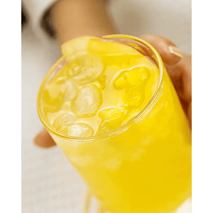 A hand holds a Thin Highball Glass (19 oz) filled with ice and a bright yellow drink, garnished with a lemon slice. Part of the Thin Highball Glasses Set of 4.