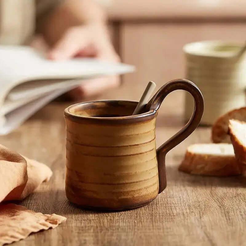 A Vintage Stoneware Espresso Cup (8–9 oz) with a spoon rests on a wooden table near sliced bread, a beige napkin, and an open book—making it the ideal unique gift for any book lover.