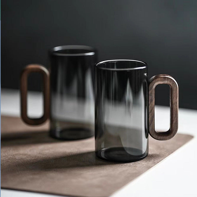 Two Macchiaco Household Borosilicate Glass Smoky Gray Water Cups with large rectangular wooden handles rest on a brown mat. These minimalist, contemporary mugs are crafted from high-quality borosilicate glass.