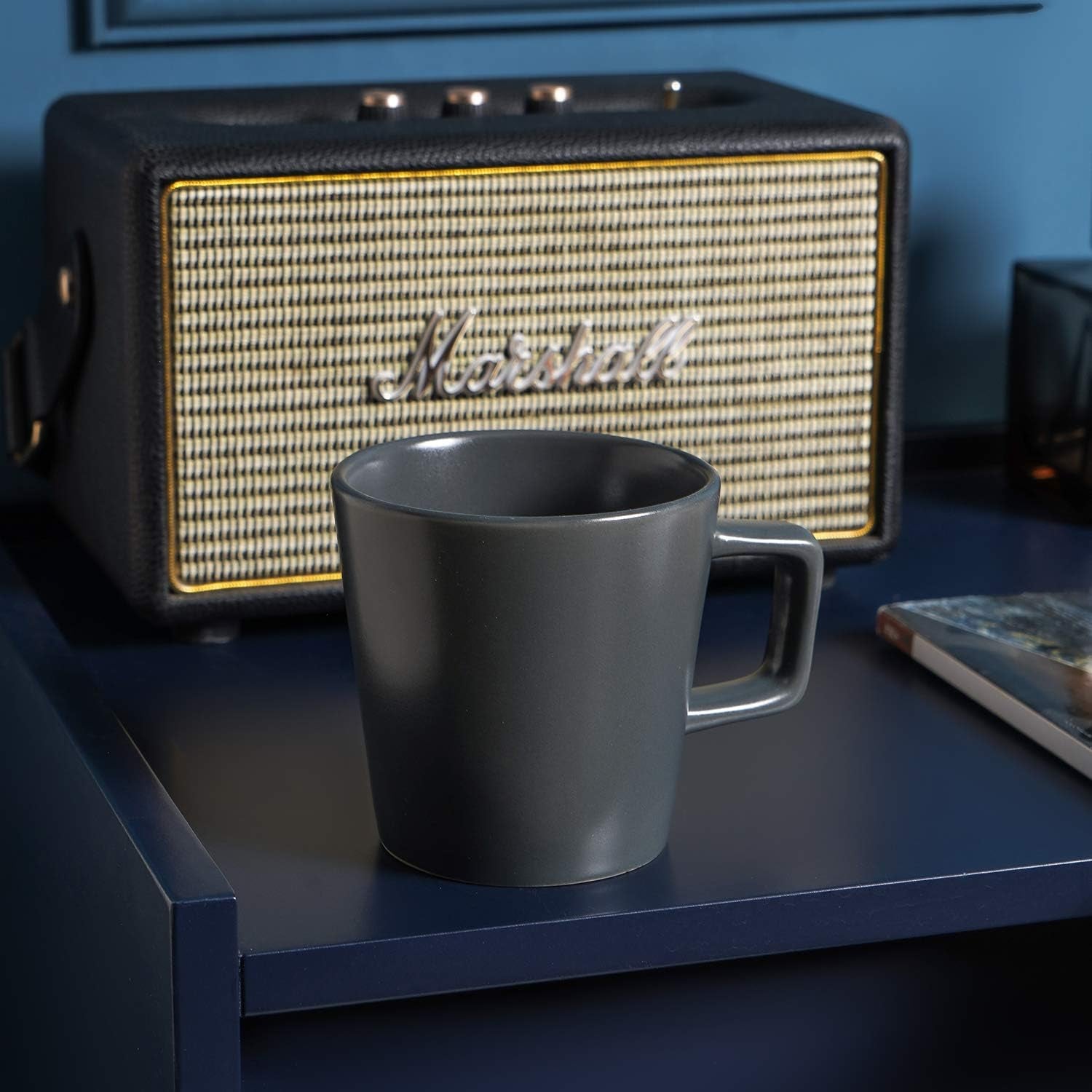 A grey matte Stoneware Coupe 12oz mug from the 4-piece set rests on a blue table in front of a Marshall speaker.