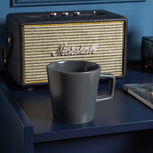 A grey matte Stoneware Coupe 12oz mug from the 4-piece set rests on a blue table in front of a Marshall speaker.