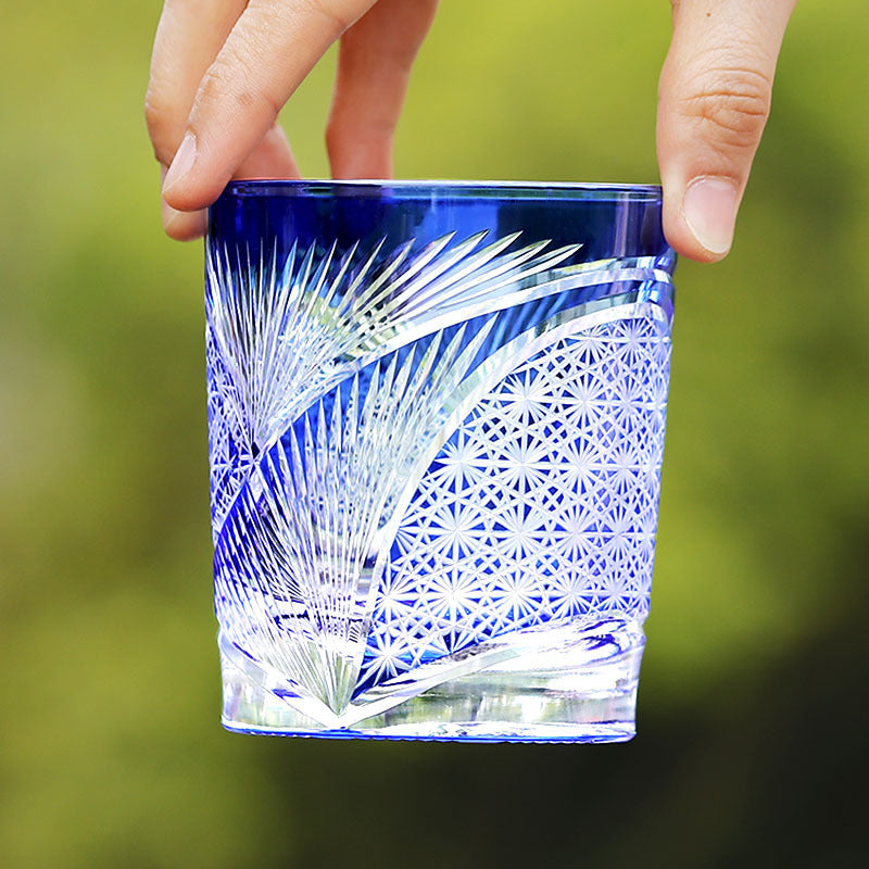 A hand holds the Macchiaco Amphora Hand-carved Whiskey Glass 8oz, featuring intricate cut geometric star patterns—an exquisite showcase of Japanese craftsmanship—set against a blurred green background.