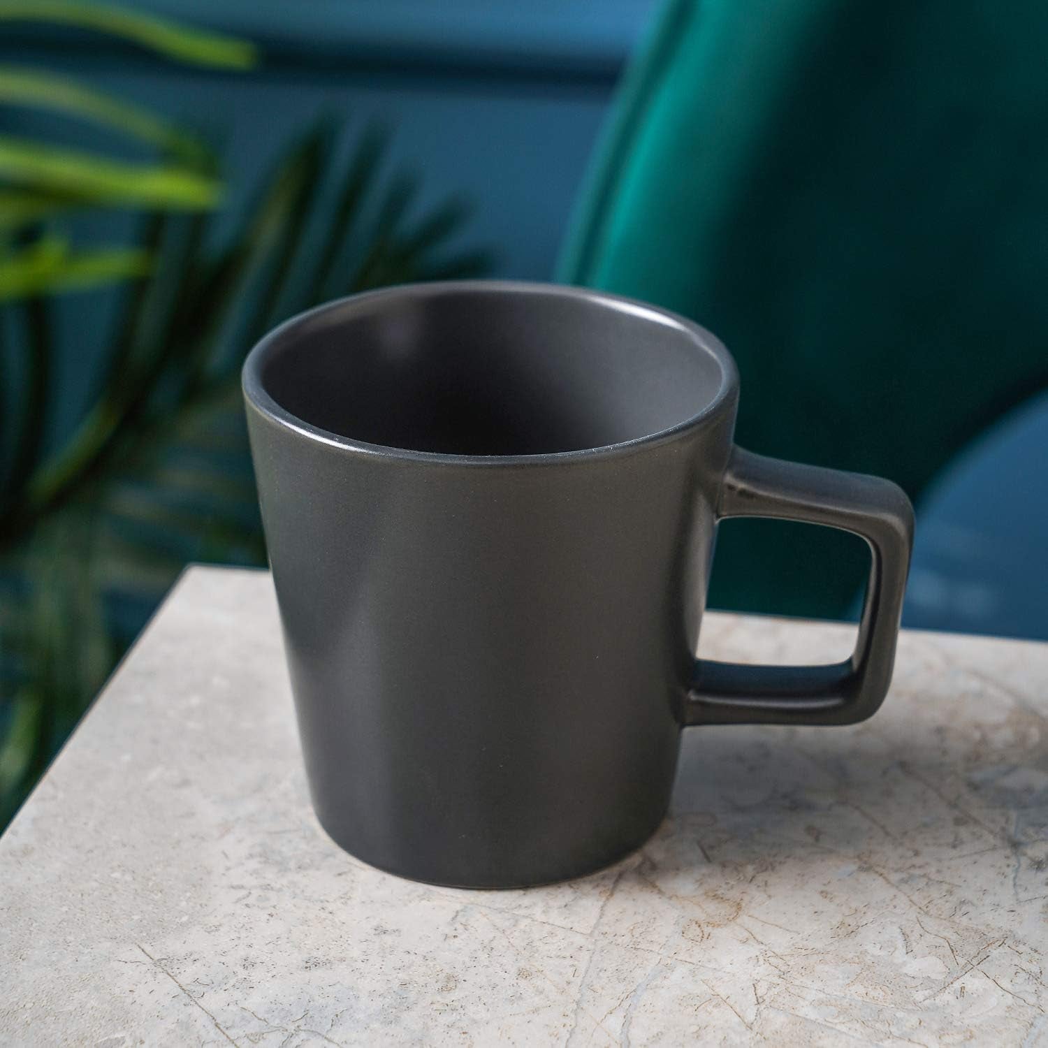 A Stoneware Coupe Mugs Set (4-piece, grey matte, 12oz) with square handles is displayed on a light marble surface, with green foliage in the background.