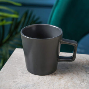 A Stoneware Coupe Mugs Set (4-piece, grey matte, 12oz) with square handles is displayed on a light marble surface, with green foliage in the background.