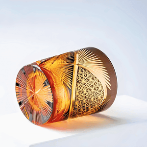 The Amphora Hand-carved Whiskey Glass 8oz features intricate orange, yellow, and gold geometric patterns, showcasing exquisite Japanese craftsmanship as it rests on its side against a plain background.