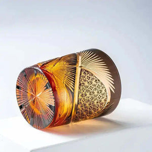 A decorative amber and gold glass tumbler with intricate geometric and fan patterns lies on its side against a plain white background.