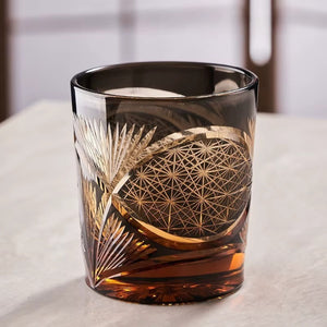 The Macchiaco Amphora Hand-carved Whiskey Glass 8oz displays Japanese craftsmanship with gold geometric and leaf patterns, resting on a light surface and partially filled with dark amber whiskey.