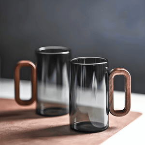 The Fiorella Smoky Fashioned Drinking Glass Set of 2 (11oz) features minimalist glass mugs with large rectangular wooden handles, resting on a brown surface against a dark, blurred background.