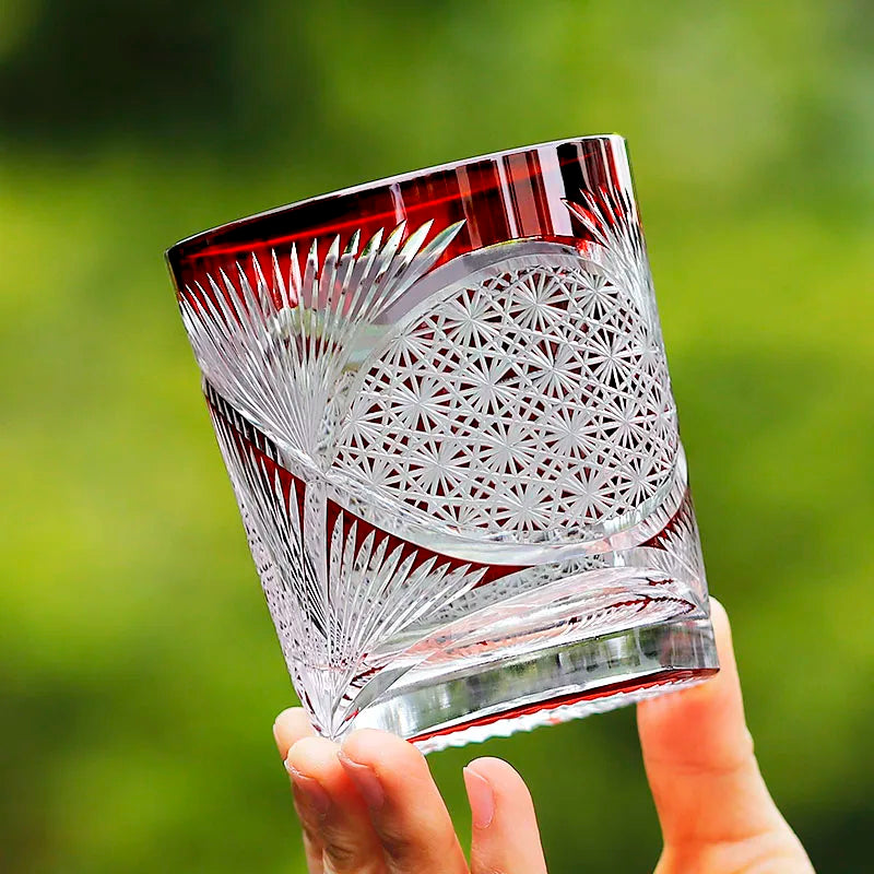 A hand holds the Macchiaco Amphora Hand-carved Whiskey Glass 8oz, showcasing etched geometric and floral patterns with a deep red rim, set against a blurred green background.