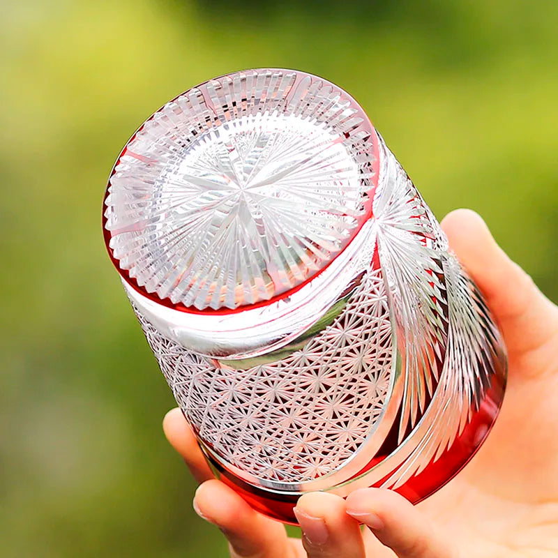 A hand holds the Macchiaco Amphora Hand-carved Whiskey Glass 8oz, adorned with red accents and Japanese-inspired geometric and floral patterns, set against a blurred green background.