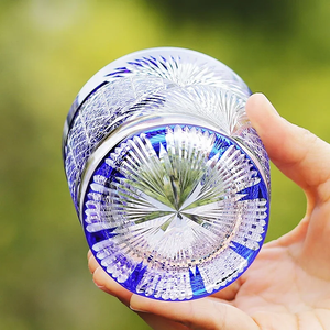 A hand holds the Macchiaco Amphora Hand-carved Whiskey Glass 8oz, featuring intricate cut patterns and starburst designs that highlight Japanese craftsmanship, set against a softly blurred green background.