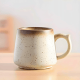 The Macchiaco 11 Oz Cocoa Coffee Mug, featuring a wide base and rounded handle with a speckled design, rests on a light wooden surface against a neutral background.