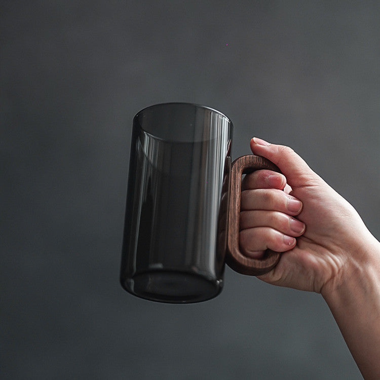 A hand holds the Fiorella Smoky Fashioned Drinking Glass (11oz, set of 2), featuring a transparent black finish and large wooden handle with minimalist design, set against a plain dark background.