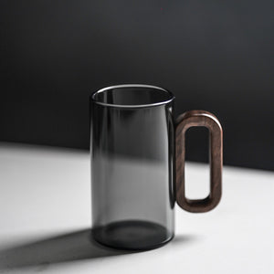 The Fiorella Smoky Fashioned Drinking Glass Set includes two 11oz transparent, cylindrical mugs featuring large oval wooden handles and a minimalist design, displayed on a white surface with a dark, blurred background.