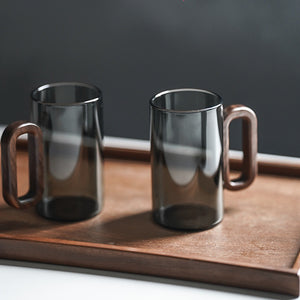 The Fiorella Smoky Fashioned Drinking Glass Set includes two 11oz transparent mugs with wooden handles, displayed side by side on a rectangular tray, highlighting minimalist elegance against a dark background.